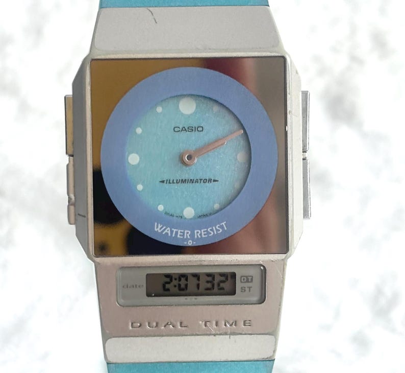 May include: A Casio dual time watch featuring a square silver-tone case with a light blue circular dial, analog hands, and a digital display window at the bottom. The watch face includes the text Casio, Illuminator, Water Resist, and Dual Time.