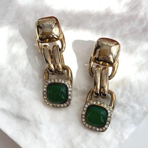 Puede incluir: Un par de pendientes dorados con una piedra central cuadrada de color verde esmeralda. Los pendientes tienen un diseño de eslabones de cadena y están incrustados con pequeños cristales transparentes alrededor de la piedra verde. Los pendientes se muestran sobre una superficie blanca texturizada.