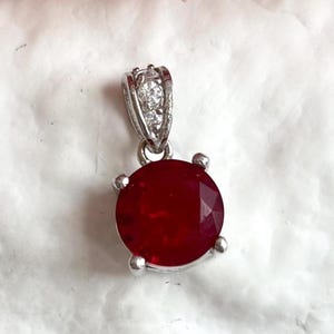 May include: A round, faceted deep red gemstone pendant set in a silver-toned metal four-prong setting. The bail is decorated with two small, clear, sparkling stones, providing a contrast to the vibrant red center stone of this jewelry piece.