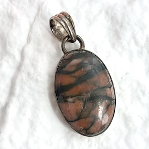 May include: Oval pendant with a silver-toned bail and a stone with a brown and black pattern. The stone is set in a silver-toned metal frame. The pendant is displayed on a white, textured surface.