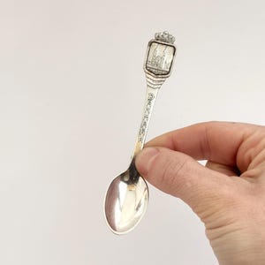 May include: A silver souvenir spoon with a rectangular emblem featuring a building and a crown. The word "DENMARK" is engraved on the handle. The spoon is held against a white background.