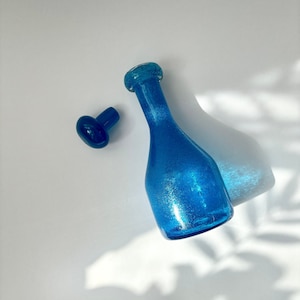 Peut inclure: Une bouteille en verre bleu avec une surface texturée et un bouchon en verre bleu assorti. La bouteille a un corps rond et un long col.