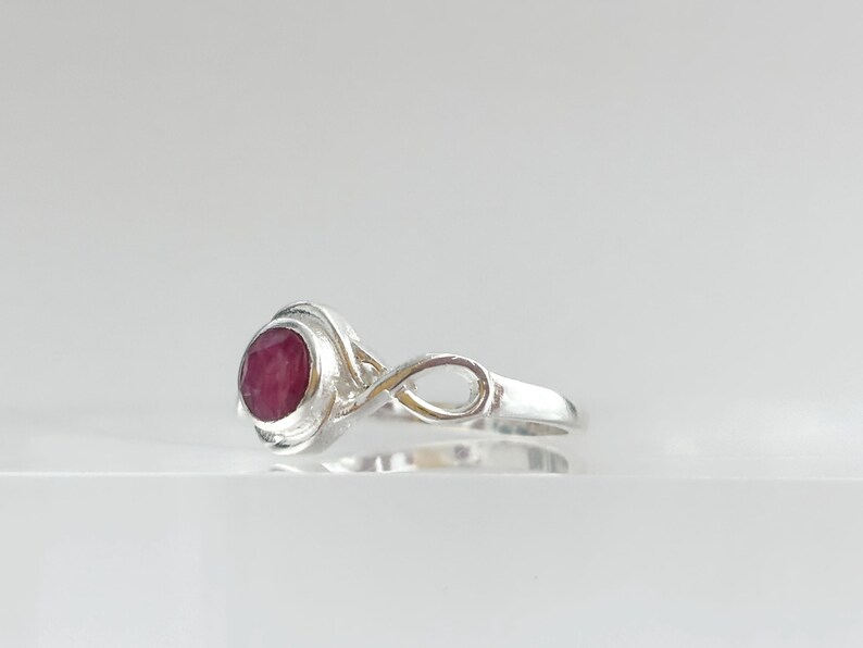 K&ouml;nnte beinhalten: Ein Silberring mit einem runden, facettierten, tiefroten Rubin, der in einer polierten Metallfassung sitzt. Die Ringschiene weist ein Unendlichkeitssymbol-Design auf, das mit der zentralen Steinfassung verbunden ist und eine Schleife bildet.