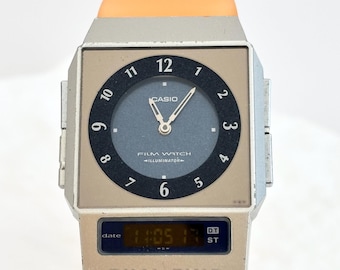 Może przedstawiać: Zegarek Casio Film Watch Illuminator z kwadratową srebrną kopertą i pomarańczowym paskiem. Tarcza posiada ciemnoniebieski okrąg z czarnymi cyframi, srebrnymi wskazówkami oraz cyfrowy wyświetlacz na dole pokazujący datę i godzinę.
