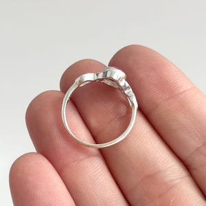 K&ouml;nnte beinhalten: Ein silberner Ring mit einem kleinen, runden, rosafarbenen Edelstein, der in eine polierte Metallschiene gefasst ist. Die Ringschiene hat eine glatte, kreisf&ouml;rmige Form mit einer dekorativen Fassung an der Oberseite, die den Stein h&auml;lt.