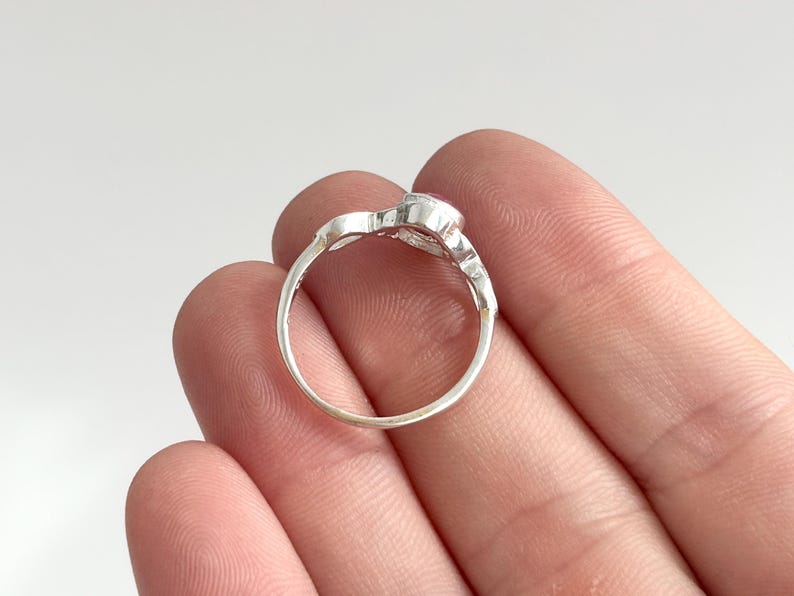 K&ouml;nnte beinhalten: Ein silberner Ring mit einem kleinen, runden, rosafarbenen Edelstein, der in eine polierte Metallschiene gefasst ist. Die Ringschiene hat eine glatte, kreisf&ouml;rmige Form mit einer dekorativen Fassung an der Oberseite, die den Stein h&auml;lt.
