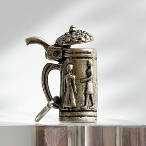 May include: A silver-toned miniature beer stein charm with an open lid. The stein features a handle and a detailed design of two figures. The lid is adorned with a textured pattern. A small, attached tag hangs from the handle.