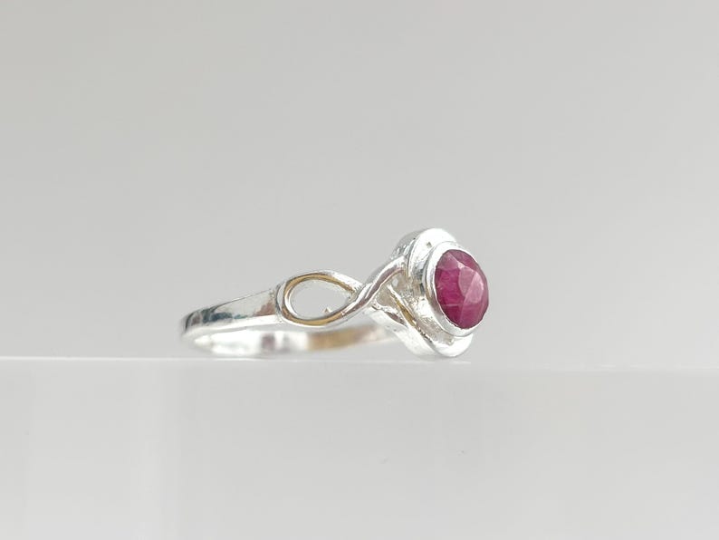K&ouml;nnte beinhalten: Ein Silberring mit einem runden, facettierten, tiefrosa Edelstein in einer Zargenfassung. Die Ringschiene zeigt ein filigranes, durchbrochenes Unendlichkeits-Schleifendesign, das mit der zentralen Steinfassung verbunden ist und eine polierte Optik bietet.