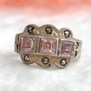 May include: A silver-toned ring featuring three square, light purple gemstones. The ring has a decorative, scalloped design with small, dark gray accents. The gemstones are set in a row, creating a vintage or antique aesthetic.