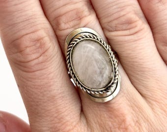 Könnte beinhalten: Silberfarbener Ring mit einem ovalen, durchscheinenden Edelstein. Der Edelstein ist in einem gedrehten, seilartigen Rand gefasst, der von einem doppelten Silberband umgeben ist. Der Ring wird an einem Finger gezeigt.