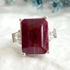 May include: A silver ring featuring a large, rectangular, deep red ruby gemstone set in four prongs. The center stone is flanked on each side by a smaller, clear, trapezoidal-cut diamond or gemstone, all mounted on a polished silver band.