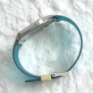 May include: A digital wristwatch featuring a silver-toned rectangular case and a smooth, light blue silicone strap. The strap is secured with a small, off-white rectangular keeper loop and a matching silver-toned metal buckle mechanism.