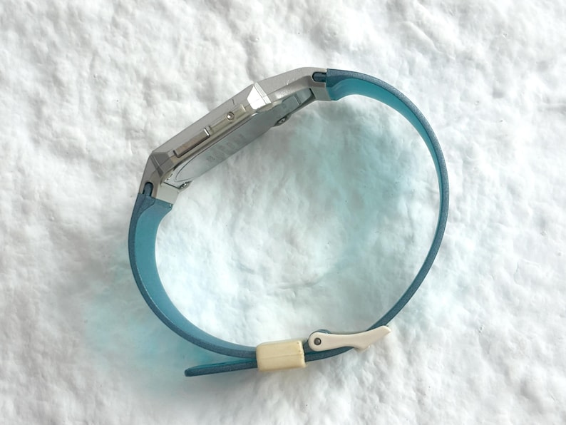 May include: A digital wristwatch featuring a silver-toned rectangular case and a smooth, light blue silicone strap. The strap is secured with a small, off-white rectangular keeper loop and a matching silver-toned metal buckle mechanism.