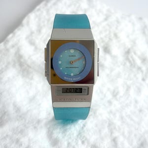 May include: A Casio wristwatch featuring a square silver-tone case with a light blue circular analog dial, gold-tone hands, and a digital display window below. The watch includes a translucent light blue silicone strap and the text DUAL TIME.