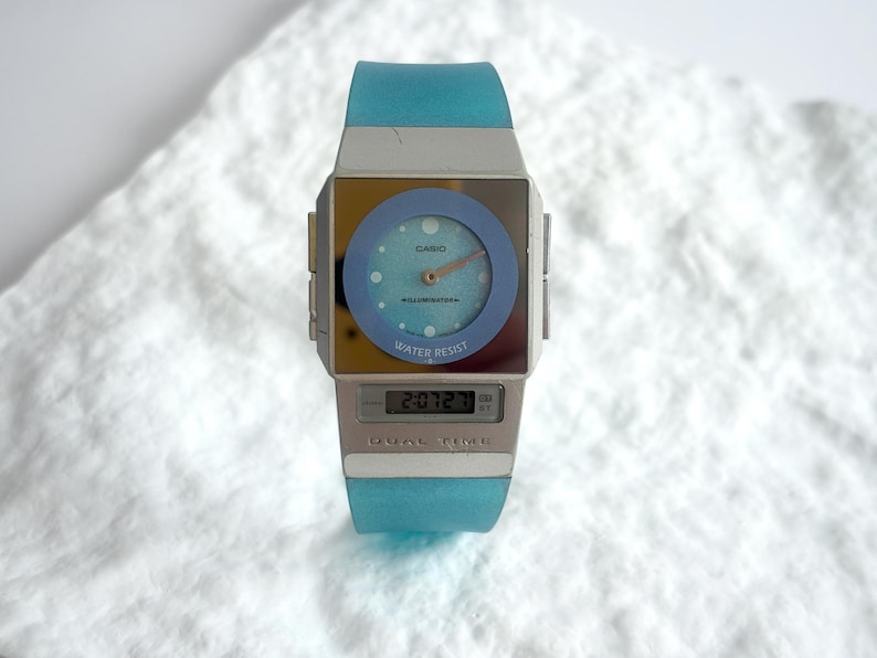 May include: A Casio wristwatch featuring a square silver-tone case with a light blue circular analog dial, gold-tone hands, and a digital display window below. The watch includes a translucent light blue silicone strap and the text DUAL TIME.
