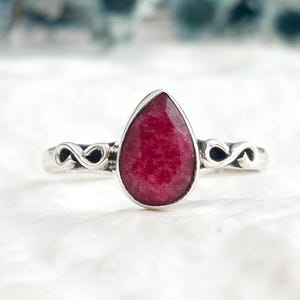 Puede incluir: Un anillo de plata con una piedra preciosa central en forma de lágrima de color rojo rosado intenso. La banda de metal presenta un diseño simétrico de bucles infinitos abiertos a ambos lados de la piedra, creando un aspecto metálico pulido.