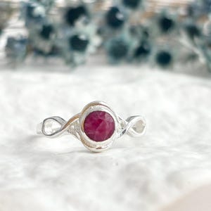 K&ouml;nnte beinhalten: Ein Silberring mit einem runden, facettierten, tief rosa-roten Edelstein in einer glatten Fassung. Die Ringschiene zeigt auf beiden Seiten des zentralen Steins ein filigranes, durchbrochenes Unendlichkeitsmuster, das ein flie&szlig;endes Design bildet.