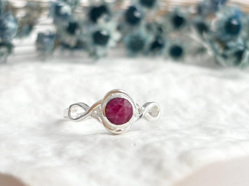 K&ouml;nnte beinhalten: Ein Silberring mit einem runden, facettierten, tief rosa-roten Edelstein in einer glatten Fassung. Die Ringschiene zeigt auf beiden Seiten des zentralen Steins ein filigranes, durchbrochenes Unendlichkeitsmuster, das ein flie&szlig;endes Design bildet.