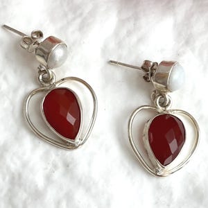 May include: A pair of silver drop earrings featuring a faceted, pear-shaped red stone set within a silver heart-shaped frame. Each earring is topped with a small, round, white stone stud, secured with a silver post and butterfly back closure.