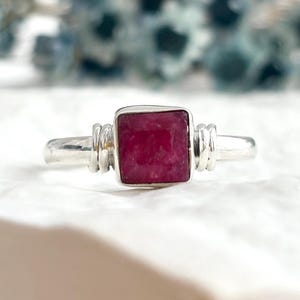 May include: A silver ring featuring a square-cut, deep pinkish-red gemstone set in a smooth metal bezel. The band is accented on both sides of the central stone with a decorative, multi-layered wire-wrapped design that adds texture to the metal.