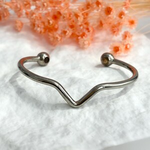 Puede incluir: Una pulsera de puño de metal plateado con un diseño en forma de V. La pulsera tiene un acabado liso y pulido y presenta pequeños acentos redondos de bola en cada extremo. La pulsera se muestra sobre una superficie blanca texturizada con flores naranjas en el fondo.
