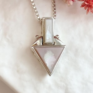 May include: A silver pendant necklace featuring a triangular pink stone with a white center inlay and a rectangular white stone accent at the top. The pendant hangs from a silver box chain, showcasing a polished metal frame and geometric design.