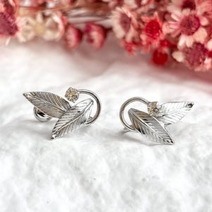 Puede incluir: Un par de pendientes plateados con forma de hoja. Cada pendiente presenta dos hojas detalladas y una pequeña gema transparente. Los pendientes están sobre un fondo blanco suave con flores borrosas rosas y rojas.
