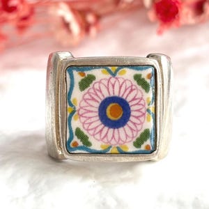 May include: A silver ring featuring a square ceramic inlay. The inlay displays a central pink flower motif with a blue and yellow center, surrounded by green and blue geometric patterns on a white background, set within a polished silver band.