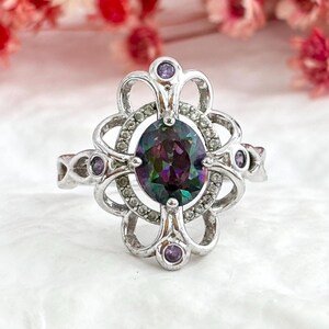 May include: A silver-toned ring featuring an oval-shaped iridescent multicolored center stone. The setting is surrounded by a halo of small clear stones and four small purple circular stones set within an intricate, openwork metal frame.