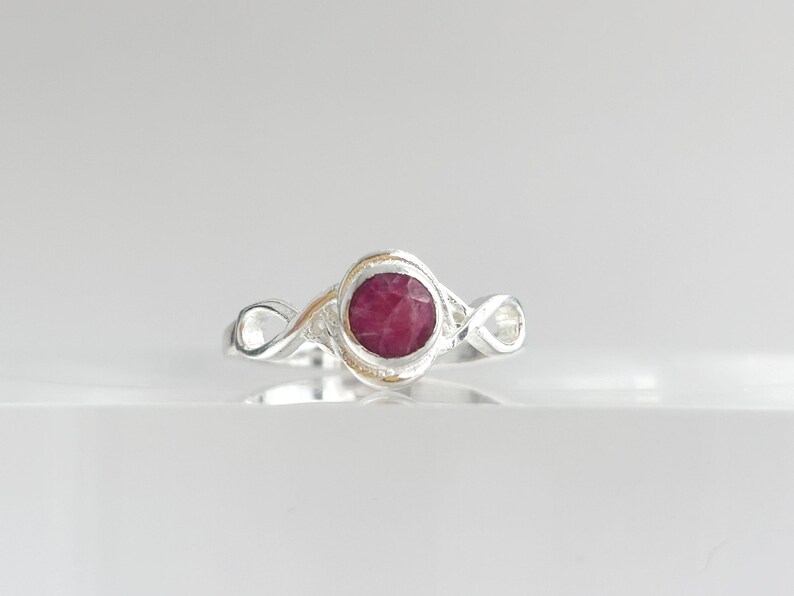 K&ouml;nnte beinhalten: Ein Silberring mit einem runden, facettierten, tief rosa-roten Edelstein in der Mitte. Die Ringschiene ist auf beiden Seiten der zentralen Fassung mit einem durchgehenden Unendlichkeits-Schleifendesign gestaltet und hat ein poliertes Finish.