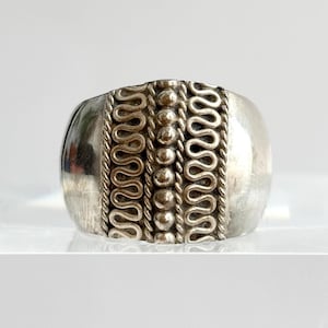 May include: A silver ring with a wide band. The ring features a central design of vertical rows of small beads and wavy lines. The ring has a polished, reflective surface and a rounded shape.