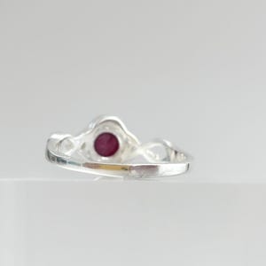 K&ouml;nnte beinhalten: Ein silberner Ring mit einem runden, tiefroten Edelstein in der Mitte. Die Ringschiene hat eine polierte, reflektierende Metalloberfl&auml;che und ein gedrehtes, durchbrochenes Design an den Seiten, das zum kreisf&ouml;rmigen Steinfassung f&uuml;hrt.