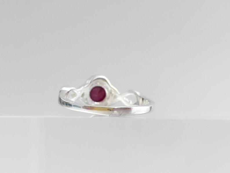 K&ouml;nnte beinhalten: Ein silberner Ring mit einem runden, tiefroten Edelstein in der Mitte. Die Ringschiene hat eine polierte, reflektierende Metalloberfl&auml;che und ein gedrehtes, durchbrochenes Design an den Seiten, das zum kreisf&ouml;rmigen Steinfassung f&uuml;hrt.