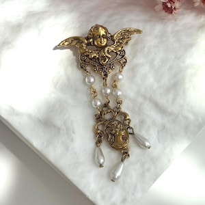 May include: A gold-toned metal brooch featuring a winged cherub at the top, connected by chains of small white faux pearls to a lower section containing an oval cameo portrait and three dangling white teardrop-shaped faux pearls.
