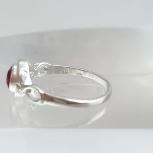 K&ouml;nnte beinhalten: Ein Silberring mit einem runden, tiefroten Edelstein in einer polierten Metallschiene. Die Ringschiene zeigt auf beiden Seiten der Fassung ein Unendlichkeits-Schleifendesign und weist einen kleinen Stempel auf der Innenseite auf.