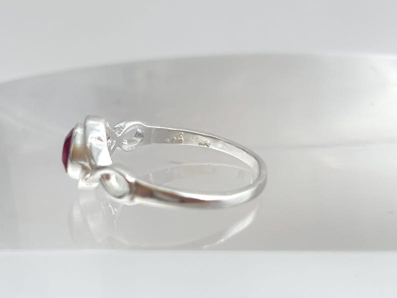 K&ouml;nnte beinhalten: Ein Silberring mit einem runden, tiefroten Edelstein in einer polierten Metallschiene. Die Ringschiene zeigt auf beiden Seiten der Fassung ein Unendlichkeits-Schleifendesign und weist einen kleinen Stempel auf der Innenseite auf.