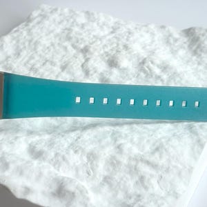 May include: A digital wristwatch featuring a metallic silver casing and a vibrant teal silicone strap. The watch face displays the text DUAL TIME in black lettering. The strap includes a series of evenly spaced rectangular adjustment holes.