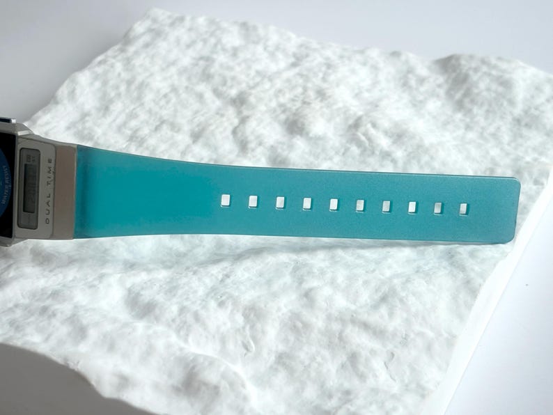May include: A digital wristwatch featuring a metallic silver casing and a vibrant teal silicone strap. The watch face displays the text DUAL TIME in black lettering. The strap includes a series of evenly spaced rectangular adjustment holes.