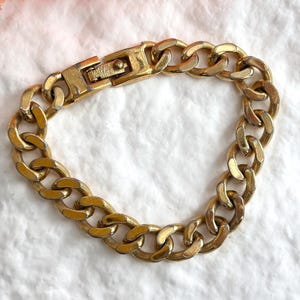 May include: A gold-toned chain bracelet with a secure clasp. The bracelet features a series of interlocking oval links, creating a bold and classic design. The metal has a slightly aged appearance, adding to its vintage appeal. The bracelet is displayed on a white textured surface.