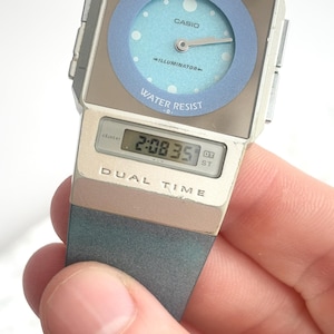 May include: A Casio Illuminator dual time watch with a silver-tone rectangular case and a light blue circular analog dial featuring white dots. The watch includes a digital display showing the time 2:08 and the text DUAL TIME on the lower case.