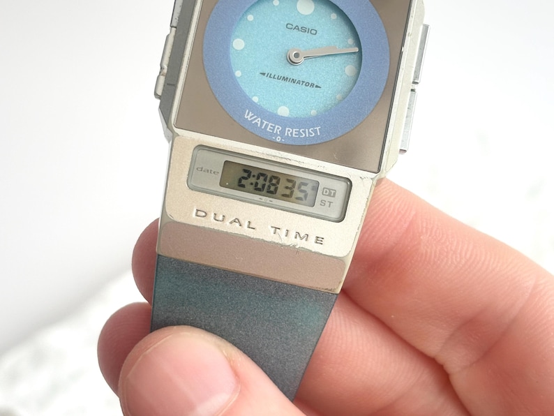 May include: A Casio Illuminator dual time watch with a silver-tone rectangular case and a light blue circular analog dial featuring white dots. The watch includes a digital display showing the time 2:08 and the text DUAL TIME on the lower case.