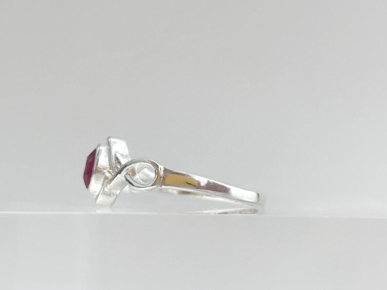 K&ouml;nnte beinhalten: Ein silberner Ring mit einem facettierten, tiefroten Edelstein in einer polierten Metallschiene. Die Schiene zeigt an der Seite ein markantes Design in Form einer Unendlichkeitsschleife, das in eine glatte, zulaufende Ringschiene &uuml;bergeht.
