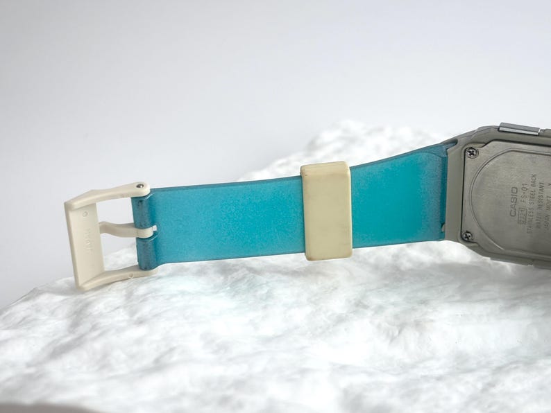 May include: A Casio watch with a translucent blue strap, a cream-colored buckle, and a cream-colored strap keeper. The stainless steel back of the watch case is visible, featuring the text Casio, FS-01, Stainless Steel Back, and Water Resistant.