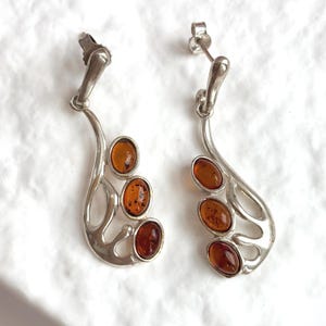 May include: A pair of silver drop earrings featuring three oval-shaped amber stones set in a curved, openwork metal frame. Each earring includes a post-back closure and a polished finish, showcasing the warm, translucent orange-brown tones.