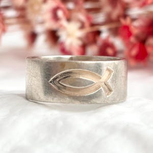 Puede incluir: Un anillo plateado con una banda ancha que presenta un símbolo Ichthys dorado. El anillo está sobre una superficie blanca y suave con un fondo borroso de flores rosas y rojas. Una pieza de joyería religiosa.