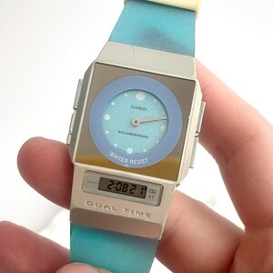 May include: A Casio Illuminator analog-digital watch with a silver-tone square case, a light blue circular dial, and a gradient turquoise silicone strap. The watch face features the text Casio, Illuminator, and Water Resist, with Dual Time below.
