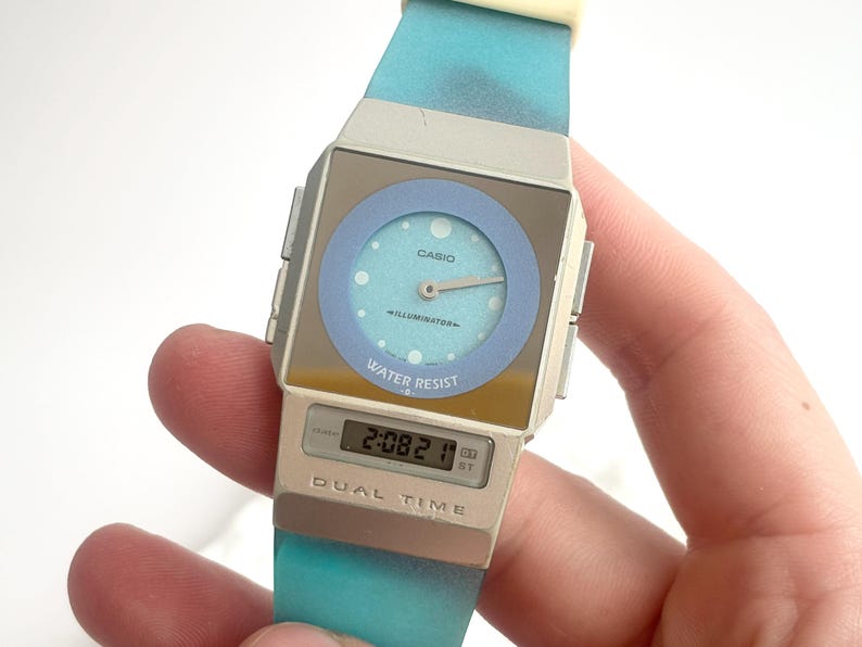 May include: A Casio Illuminator analog-digital watch with a silver-tone square case, a light blue circular dial, and a gradient turquoise silicone strap. The watch face features the text Casio, Illuminator, and Water Resist, with Dual Time below.