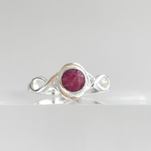 K&ouml;nnte beinhalten: Ein Silberring mit einem runden, facettierten, tief rosa-roten Edelstein in der Mitte. Die Ringschiene ist poliert und weist auf beiden Seiten der zentralen Steinfassung ein Design mit unendlich geformten Schlaufen auf.