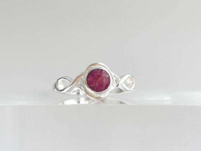 K&ouml;nnte beinhalten: Ein Silberring mit einem runden, facettierten, tief rosa-roten Edelstein in der Mitte. Die Ringschiene ist poliert und weist auf beiden Seiten der zentralen Steinfassung ein Design mit unendlich geformten Schlaufen auf.