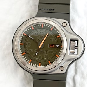 Puede incluir: Un reloj de pulsera con esfera verde, marcadores naranjas y bisel plateado. El reloj tiene una correa verde oscuro y una pantalla de fecha a las 3 en punto. La esfera del reloj tiene el texto "AUTOMATIC 20 JEWELS". El reloj es de estilo militar.